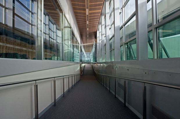 A long enclosed commercial interior passage or ramp with accessible handrails, dark carpet flooring, extensive floor-to-ceiling glazing on both sides, exposed structural framing, and a warm timber slatted ceiling. The corridor connects contemporary institutional or office spaces and is brightly illuminated by natural light.