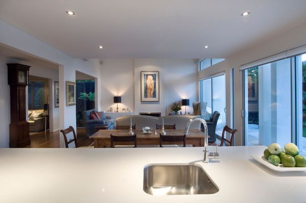 Open-plan interior viewed from a kitchen island, with a polished light benchtop and undermount stainless-steel sink in the foreground. Beyond it is a dining area with a timber table and chairs, opening into a bright contemporary living space with sofas, armchairs, artwork, recessed ceiling lights, and large floor-to-ceiling glazed doors and windows.
