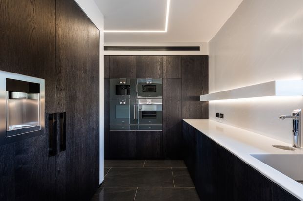 A sleek contemporary galley kitchen with tall dark timber-look cabinetry, integrated black appliances, a long white stone benchtop, undermount sink and minimalist chrome tapware. Large dark-format floor tiles and concealed linear LED lighting create a refined, high-contrast interior.