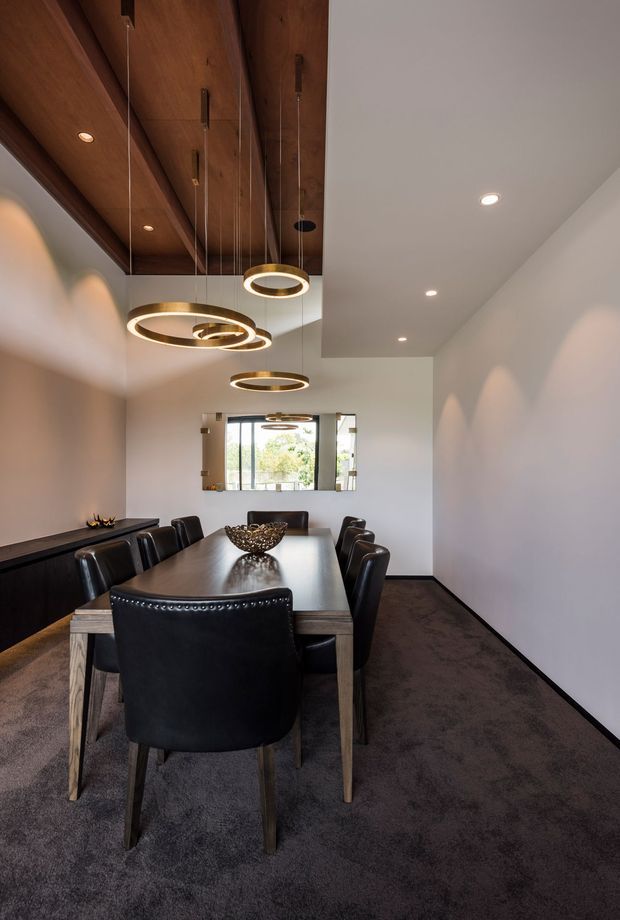 A contemporary dining room with a long rectangular timber dining table surrounded by black leather chairs. The room has dark carpet flooring, white walls, a dramatic timber-lined ceiling panel, recessed downlights, and multiple suspended circular pendant lights. A window at the far end brings in natural light and overlooks greenery.