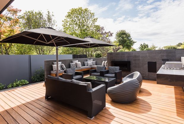 A spacious contemporary outdoor entertaining deck with warm timber decking, dark built-in masonry walls, a large outdoor fireplace, and a sleek outdoor kitchen or bar. Black outdoor sofas and armchairs are arranged around a low coffee table beneath two large cantilevered umbrellas, with sculptural seating and landscaped greenery around the perimeter.