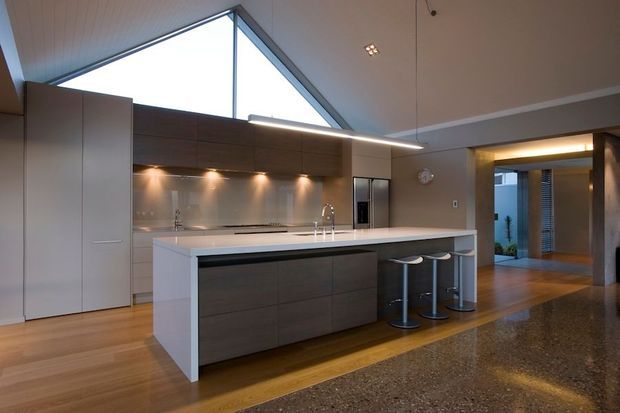 A contemporary open-plan kitchen with a large island, integrated dark timber cabinetry, pale benchtops, stainless steel appliances, and three bar stools. A dramatic triangular clerestory window brings in natural light, while a polished concrete floor contrasts with timber flooring in the kitchen zone. The room opens toward a hallway or entry area.