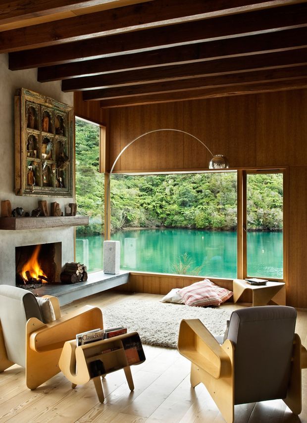 Warm, mid-century-inspired living room with a dramatic exposed timber-beam ceiling, rich timber wall panelling, and expansive corner glazing overlooking a turquoise lake and dense greenery. A masonry fireplace with an active fire anchors the room, complemented by sculptural wood-and-leather lounge chairs, a low magazine rack, a pale shag rug, and a built-in window seat.