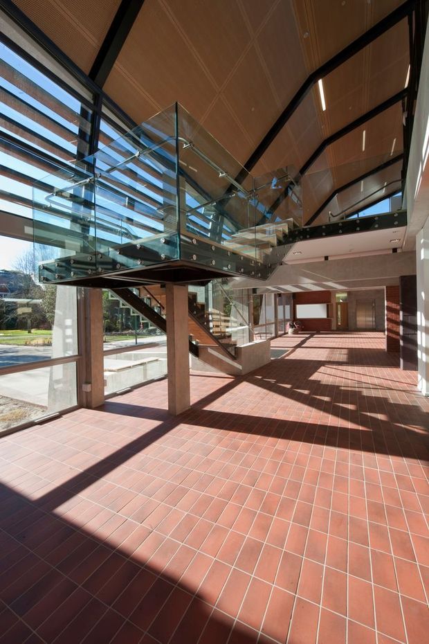 A spacious, sunlit institutional or commercial interior with a dramatic suspended glass stair and landing. The space features extensive floor-to-ceiling glazing, exposed dark structural framing, warm timber-lined vaulted ceilings, terracotta square tile flooring, timber columns, and strong geometric shadows. A landscaped exterior is visible through the glazing.