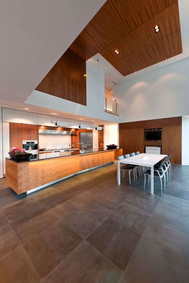 A spacious contemporary open-plan kitchen and dining area with a dramatic double-height ceiling. The kitchen features extensive dark timber cabinetry, a large timber island with stainless-steel work surfaces, integrated appliances, and a pale reflective splashback. A white dining table with multiple chairs sits adjacent to the kitchen. Large-format dark grey floor tiles, white walls, recessed lighting, and a timber-lined ceiling create a warm, refined modern interior.