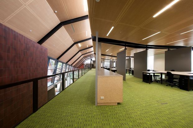 Open-plan commercial office interior with a long circulation gallery and workstations. The space features a dramatic sloping timber-lined ceiling supported by exposed black structural beams, a red-brown tiled feature wall, extensive glazing along one side, green carpet flooring, and freestanding white and timber-toned partitions and desks.
