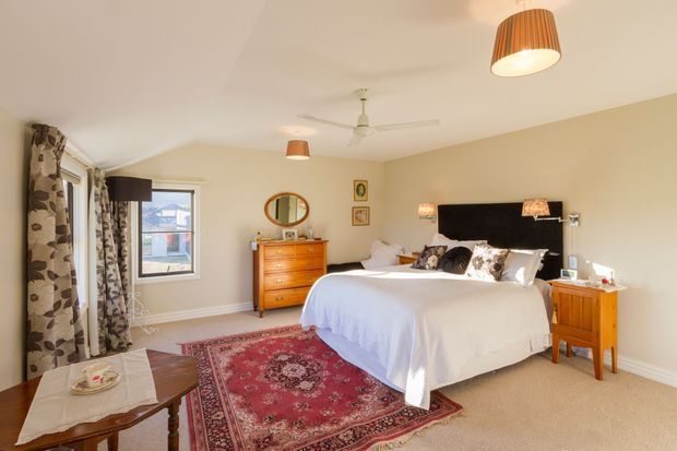 A bright, warmly styled bedroom with a large bed, black upholstered headboard, white bedding, patterned accent pillows, a red Persian-style area rug, timber chest of drawers and bedside table, patterned curtains, ceiling fan, and pendant lighting. The room has beige carpet and light cream walls, with a window overlooking neighboring buildings.