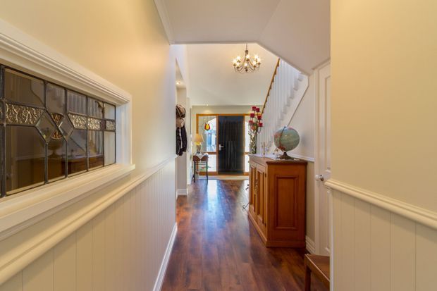 A traditional residential hallway viewed toward an entry door and staircase. The corridor features dark polished timber flooring, cream painted walls with white wainscoting and trim, a decorative leaded-glass internal window, a timber console cabinet, and a classic chandelier. The staircase rises along the right side, with an entry area visible beyond.