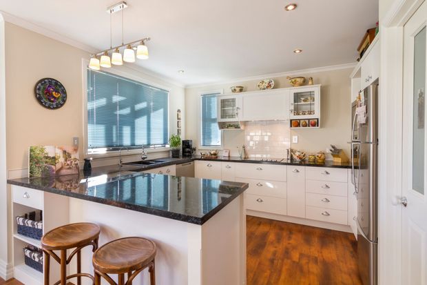 Bright, traditional-style residential kitchen with a large central island, dark polished stone benchtops, white shaker-style cabinetry, a pale tiled splashback, timber flooring, stainless-steel appliances, and pendant lighting. The room includes a small breakfast seating area with two wooden stools and ample built-in storage.