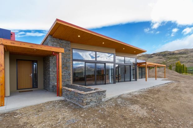 Exterior view of a contemporary single-storey mountain home with a broad projecting roof, exposed warm timber soffits, dark metal-framed floor-to-ceiling glazing, and natural stacked-stone walls. A covered entry and open-sided timber carport extend from the main volume, with rugged mountainous terrain visible behind the building.
