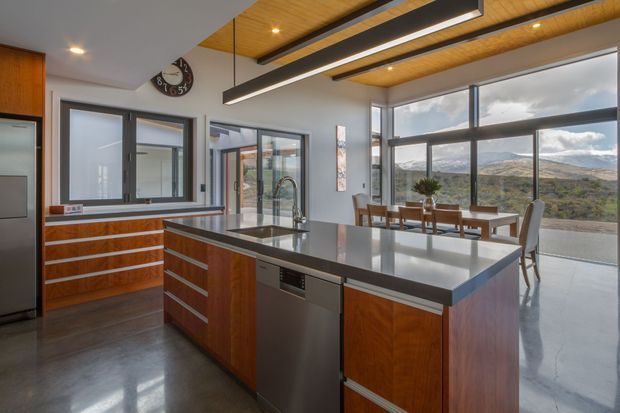 A contemporary open-plan kitchen with a large central island, dark engineered stone benchtops, warm timber cabinetry, stainless-steel appliances, and polished concrete flooring. A timber-lined ceiling with linear pendant lighting spans the space, while floor-to-ceiling glazing connects the kitchen and dining area to a broad natural landscape.