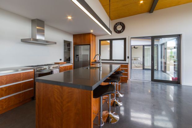 A spacious contemporary kitchen with a large dark stone island and integrated bar seating, warm timber cabinetry, stainless-steel appliances, and polished concrete flooring. The room has a partially timber-lined ceiling with exposed dark structural beams, linear recessed lighting, and extensive glazing and sliding doors opening toward an adjacent living area. A dining or living space is visible beyond the kitchen.
