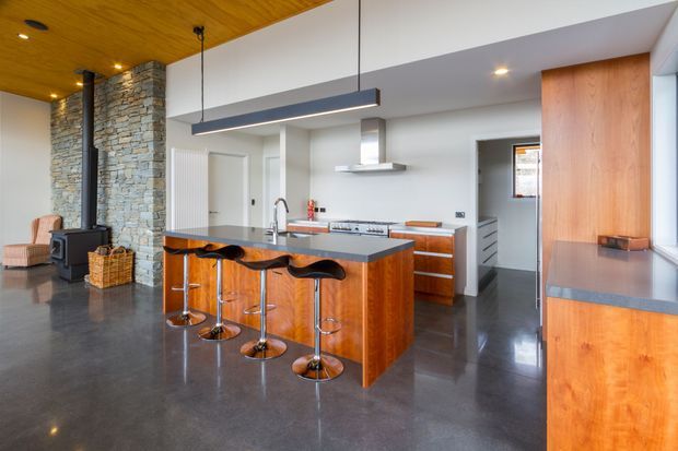 Open-plan kitchen with a large timber island, dark stone benchtops, stainless-steel appliances, polished concrete flooring, exposed stone fireplace column, and a warm timber-lined ceiling. A dining and living area is visible beyond the kitchen, with contemporary pendant lighting and integrated timber cabinetry.