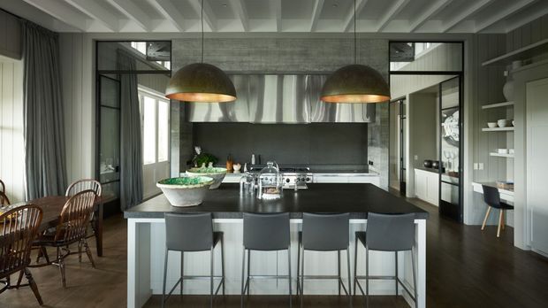 Contemporary open-plan kitchen dominated by a large dark island with four bar stools, dark cabinetry, stainless-steel splashback and appliances, and twin oversized dome pendant lights. A dining area, glazed partitions, and built-in shelving are visible around the kitchen. The ceiling features exposed painted structural beams, while timber flooring and grey cabinetry create a refined industrial-modern atmosphere.