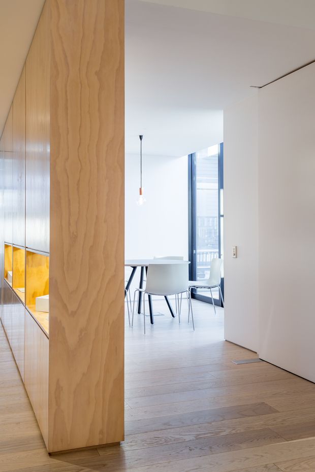 A bright, minimalist residential interior viewed through an open doorway. The foreground features a pale timber floor and a full-height timber-clad partition or built-in joinery wall with a warm illuminated niche visible at the left. Beyond it is a dining area with a white table and white chairs, floor-to-ceiling glazing, and a slim pendant light. White walls, a clean-lined ceiling, and dark window frames create a crisp contemporary composition.
