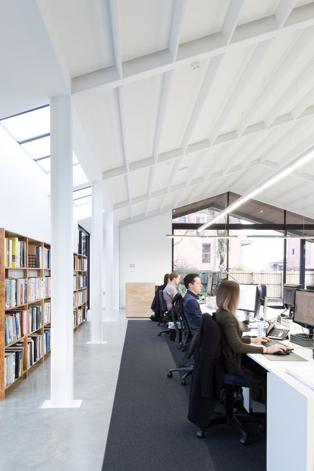 Bright open-plan commercial office interior with rows of workstations, employees using computers, extensive bookshelves, polished concrete flooring, white exposed roof trusses and columns, and large glazed openings. A timber-lined angular ceiling feature and linear pendant lighting define the workspace.