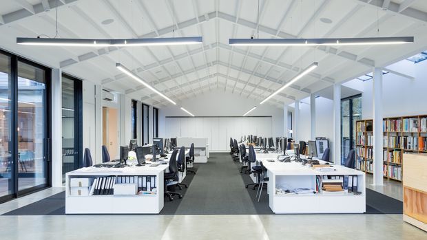 A bright contemporary open-plan office with rows of white workstations, computer monitors, task chairs, and integrated open shelving. The space has a high exposed white pitched roof structure, exposed beams and rafters, linear suspended lighting, full-height black-framed glazing, and a grey carpeted central circulation zone. Bookshelves and storage line the right side, creating a clean, airy workplace with a strong architectural ceiling presence.