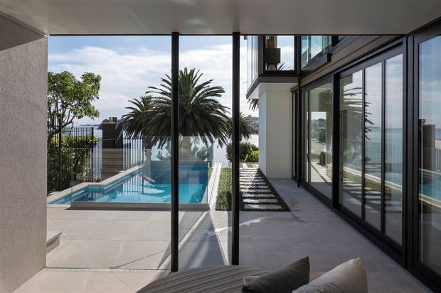 A contemporary covered outdoor terrace or poolside area beside a modern multi-level home. Full-height black-framed glazing lines the right side, reflecting the pool and surrounding landscape, while a covered ceiling and slim dark structural posts frame views toward a rectangular swimming pool, tiled deck, hedging, palm trees, and coastal water beyond. A partial outdoor sofa is visible in the foreground.