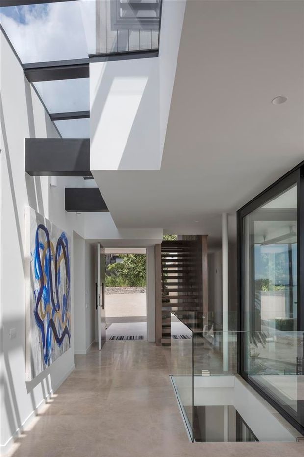 A bright, contemporary interior hallway or entry gallery with polished pale stone flooring, white walls, black-framed floor-to-ceiling glazing, and a dramatic glazed skylight or atrium above. A floating staircase with dark treads and a timber slatted screen are visible toward the rear, while an abstract blue artwork adds colour to the wall. The space has strong natural light, clean lines, and a visually open connection between levels.