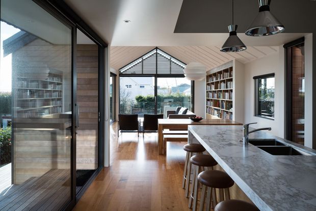 Open-plan contemporary kitchen and living area with a long marble island, dark stools, timber flooring, extensive built-in timber bookshelves, and large glazed openings. The space features a pitched, timber-lined ceiling over the living area, pendant lights, and views into a landscaped suburban setting.
