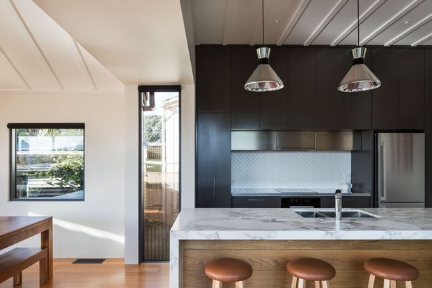 A contemporary kitchen viewed from an adjacent dining area, featuring a large white marble-look island with a timber-clad breakfast-bar face and three brown leather stools. The kitchen has dark full-height cabinetry, integrated stainless-steel appliances, a pale chevron-patterned splashback, twin metallic pendant lights, and warm timber flooring. A narrow glazed door and windows bring in natural light.