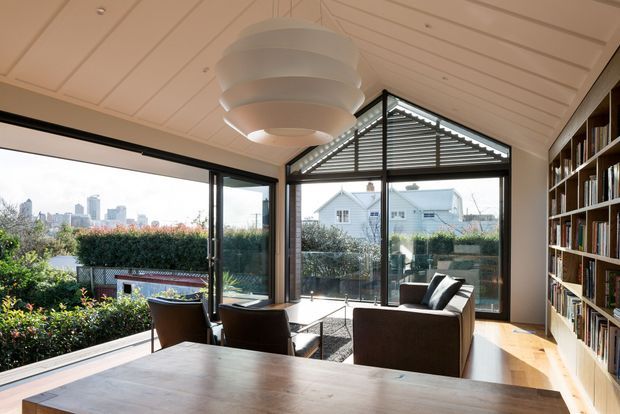 Bright, contemporary sunroom or living area with extensive black-framed glazing, a dramatic glazed gable, white slatted pitched ceiling, built-in full-height bookcase, timber flooring, and dark upholstered seating around a low table. A large sculptural pendant hangs centrally, while landscaped outdoor space and neighbouring houses are visible through the windows.