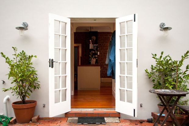 Front-facing view of a traditional home entry with white French doors opened onto a small brick-paved threshold. The entrance has smooth white rendered walls, wall-mounted exterior lights, potted citrus-like plants, brick paving, and a warm timber-floored interior with exposed brick visible beyond.