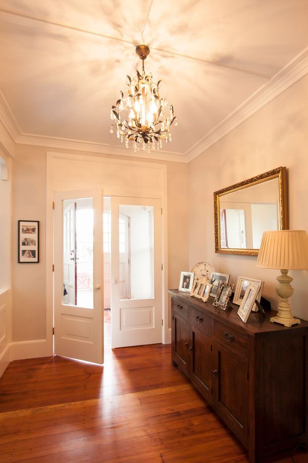 Elegant traditional entry hall with polished dark timber flooring, warm cream walls, ornate layered ceiling cornices, and white panelled glazed double doors opening to a bright exterior space. A dark timber sideboard with framed photographs sits beneath a large ornate gold-framed mirror, complemented by a pleated table lamp. A decorative crystal and metal chandelier is the central feature, creating a warm, refined atmosphere.