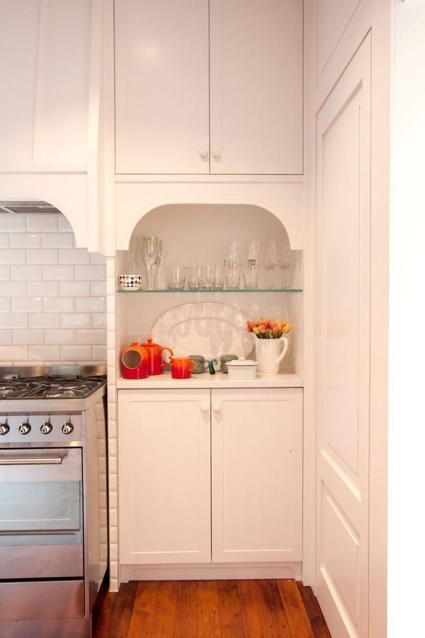 A compact, traditionally styled kitchen vignette featuring tall white built-in cabinetry with paneled doors and a recessed arched display niche. The niche has a glass shelf and displays glassware, ceramics, a white platter, and colourful red and orange serving pieces. At left is a stainless-steel range and oven beneath a white subway-tile backsplash, while the floor is finished in warm timber boards.