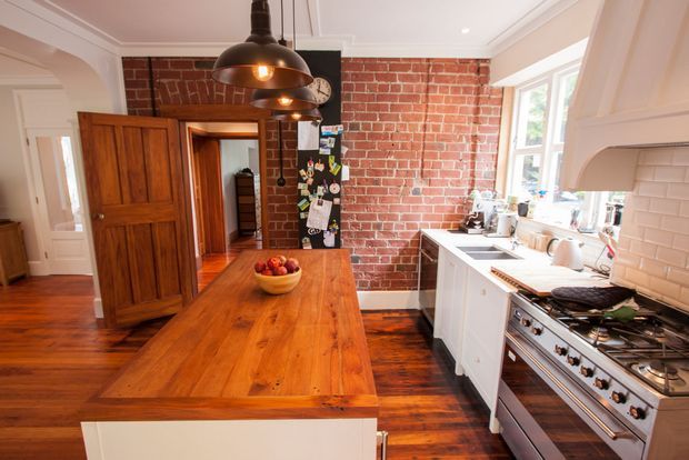 A warm, character-filled kitchen featuring a large solid timber island with seating, white cabinetry, a white subway-tile splashback, exposed red brick walls, and dark timber flooring. A stainless-steel range cooker and sink sit beneath bright windows, while black industrial pendant lights hang over the island. Timber panelled doors and traditional architectural trims are also visible.