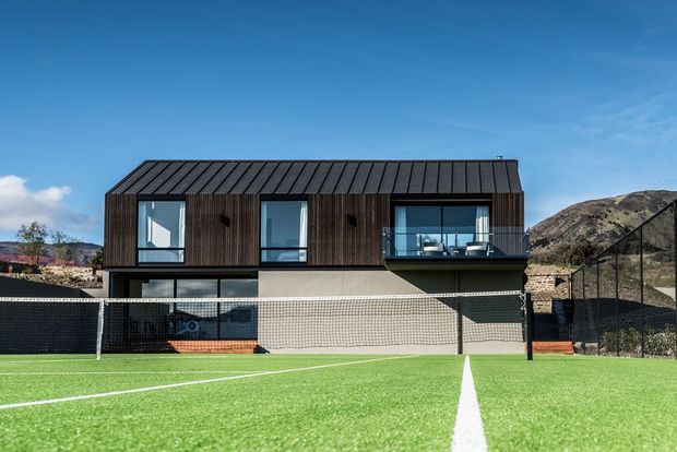 Modern two-storey rectangular house with dark vertical timber cladding, a standing-seam dark metal pitched roof, large windows and a projecting glass-fronted upper balcony. The building sits directly behind a fenced artificial-turf sports court, with a dry rocky hillside in the background.