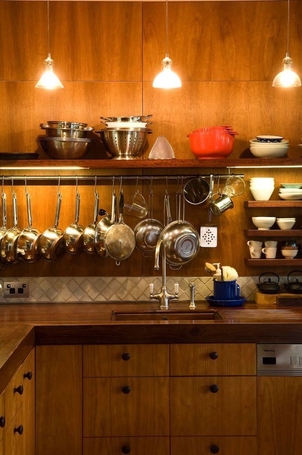 A compact, richly timber-lined kitchen with extensive open shelving and hanging rails displaying copper and stainless-steel cookware. Three pendant lights illuminate a tiled backsplash, deep timber benchtop, farmhouse-style sink with brass-toned tapware, and warm wood cabinetry with dark knobs. The space has a curated, traditional cooking-kitchen character.