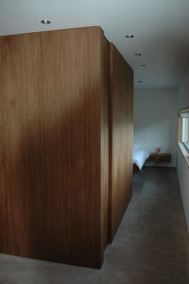 Interior view of a minimalist corridor or landing with extensive full-height dark timber wall panelling and built-in joinery. A partially visible bedroom with white bedding is at the far end, alongside a narrow horizontal window. The space has a smooth pale ceiling with recessed downlights and a grey polished concrete floor.