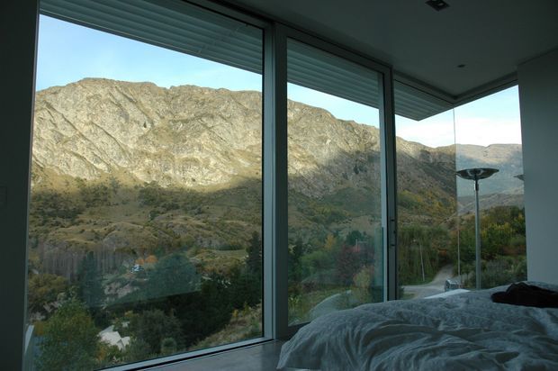 Interior bedroom view with floor-to-ceiling corner glazing overlooking a rugged mountainous landscape. A partially visible bed with white bedding occupies the foreground, while dark slim window frames, exterior roller blinds, and a concealed ceiling with recessed downlights create a restrained contemporary setting.