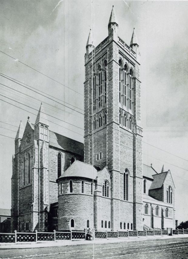 Historic black-and-white photograph of a large Gothic Revival church constructed in brick or masonry, featuring a prominent square bell tower with four pointed corner turrets, tall pointed-arch windows, steep gabled roofs, buttresses, and a rounded apse-like projection. The building is viewed from the street behind a decorative fence, with overhead utility wires and a vintage photographic appearance.