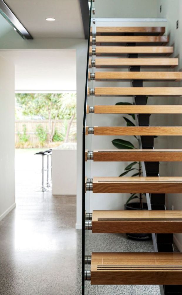 Bright contemporary interior view centred on a floating timber staircase with open-riser treads, dark structural supports, polished metal fixings and a clear glass balustrade. A light-toned hallway and adjacent space are visible, with terrazzo-like flooring, white walls, a roller blind, and indoor planting.