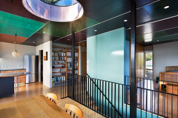 A contemporary open-plan upper-level interior combining a kitchen, dining area, library shelving and a stair landing. The space features warm timber flooring and dining furniture, black metal balustrades, extensive built-in book shelving, glossy dark ceiling panels with colourful translucent sections, and a large circular skylight. A turquoise glazed or painted feature wall overlooks the stairwell, while a timber seating area is visible beyond.