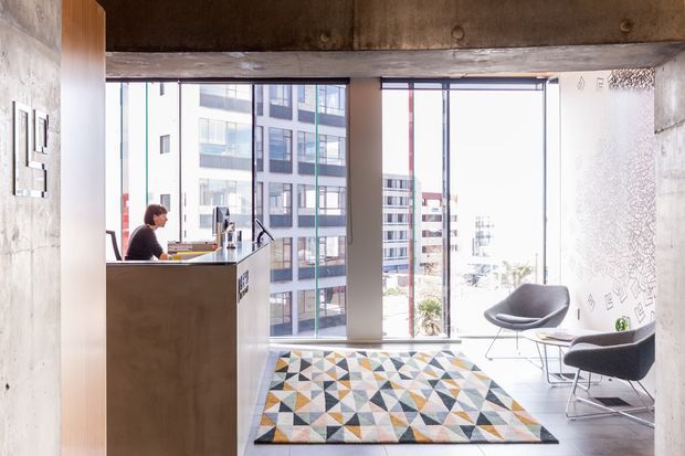 Bright contemporary commercial reception or waiting area with exposed concrete walls and ceiling, large floor-to-ceiling windows overlooking dense urban buildings, a simple timber-toned reception desk, and two modern lounge chairs around a small table. A bold geometric patterned rug adds colour to the otherwise neutral space.