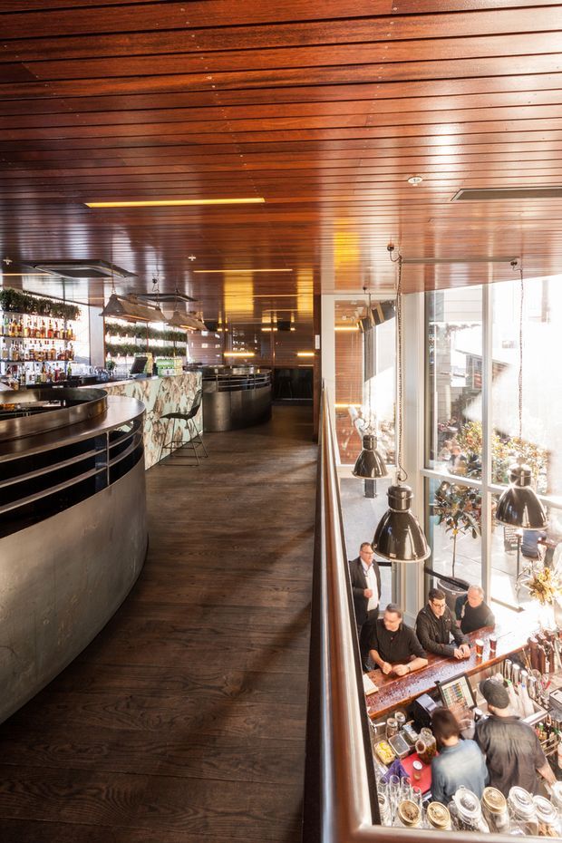 A bright, multi-level commercial bar and dining venue viewed from an upper level. The space features a long dark timber floor, a richly stained slatted timber ceiling, large windows, hanging pendant lights, curved black metal bar counters, bottle displays, and a busy bar area below. The interior has a warm, refined hospitality design with strong vertical openness between levels.