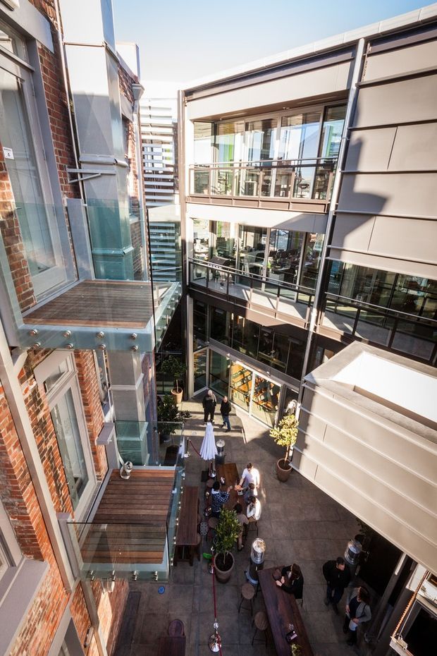 A high-angle view into a narrow multi-level urban courtyard or atrium between contemporary and adapted brick buildings. The space features extensive glazing, steel-framed balconies and walkways, timber balcony decking, horizontal metal cladding, and a paved ground-level gathering area with tables, planters, and people. Bright daylight and strong vertical perspectives emphasize the tall, enclosed commercial-style courtyard.