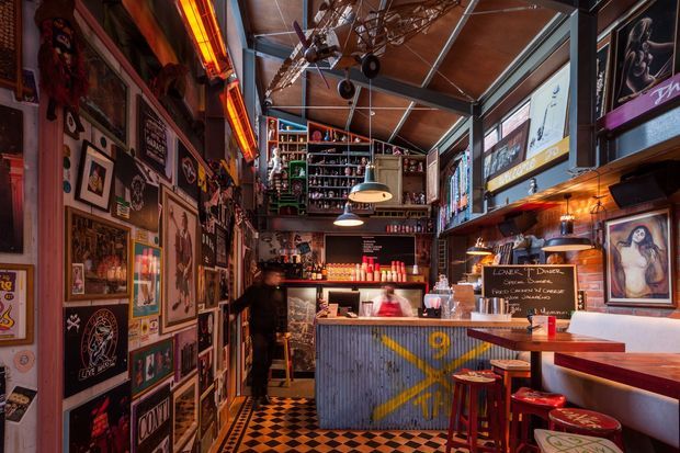 A compact, richly decorated bar or café interior with exposed structural framing, a high sloped ceiling, corrugated metal bar frontage, exposed brick walls, extensive eclectic wall art and signage, pendant lighting, stools, and a black-and-white checkerboard floor. The space has a strong vintage industrial character with bold red, orange, and metallic accents.