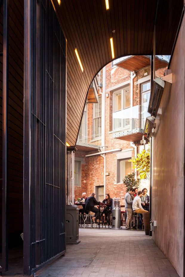 A narrow, covered urban courtyard or laneway connecting to a small outdoor dining area. The space features a dramatic curved timber-lined canopy with integrated linear lighting, dark vertical metal screening, exposed red-brick walls, glazed balcony balustrades, and paved flooring. Several patrons sit and stand around café tables at the far end, creating a lively hospitality setting.