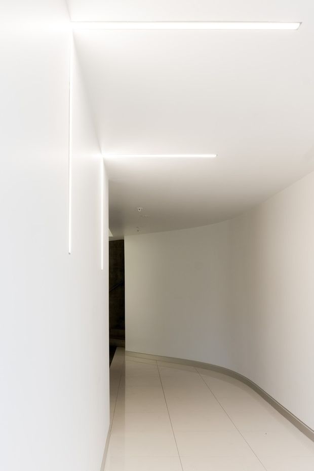A minimalist, curved interior corridor or passage with smooth white walls, a gently curved ceiling and skirting, pale large-format tiled flooring, and recessed linear LED lighting. The space is empty and clean, with a darker opening visible at the far end.
