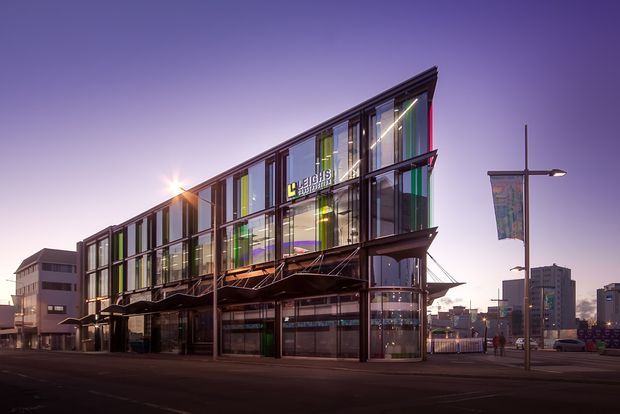 Exterior view of a contemporary multi-storey commercial building at dusk. The building has an angular glazed facade with extensive floor-to-ceiling windows, dark metal framing, colourful vertical panels, projecting structural elements, and prominent signage. It occupies an urban streetscape with street lighting and neighbouring commercial buildings.