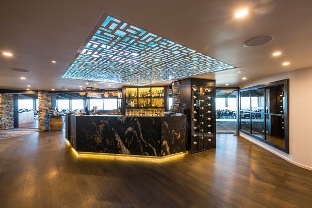 A sophisticated interior bar and lounge area dominated by a large black marble-clad bar island with warm underlighting. Behind it is a back bar with illuminated bottle displays and glass-fronted wine storage. The room has dark timber flooring, exposed stone feature walls, extensive glazing, and a striking geometric illuminated ceiling feature in blue-white tones. Dining and lounge areas are visible through the openings and glass partitions.