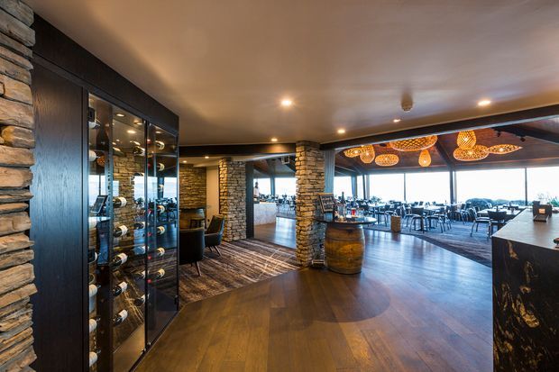 A refined commercial dining and lounge interior with a wine display wall, dark timber flooring, stacked-stone columns and walls, exposed dark ceiling beams, and warm recessed lighting. The space opens into a bright dining area with extensive glazing, contemporary tables and chairs, and distinctive woven pendant lights. A barrel and dark stone service counter reinforce the restaurant or hospitality setting.