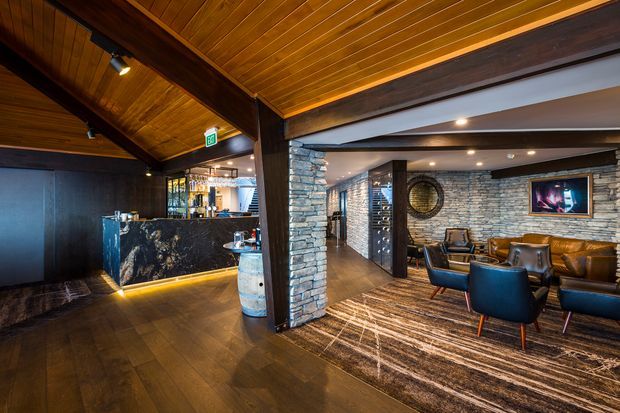 A sophisticated commercial lounge and bar interior with dark timber flooring, a richly timber-lined ceiling, exposed dark structural beams, and a dramatic black stone bar counter. The adjoining seating area features stacked natural-stone walls, leather lounge seating, a large patterned rug, recessed lighting, and a wall-mounted screen. The atmosphere is warm, moody, and upscale.