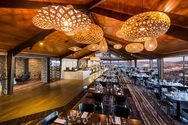 A spacious upscale restaurant interior viewed from the dining area, with extensive tables and chairs, a long bar and service counter, dramatic woven pendant lights, dark timber ceilings and floors, exposed structural beams, and floor-to-ceiling glazing overlooking an urban landscape. A stacked-stone feature wall and warm ambient lighting add texture and richness.