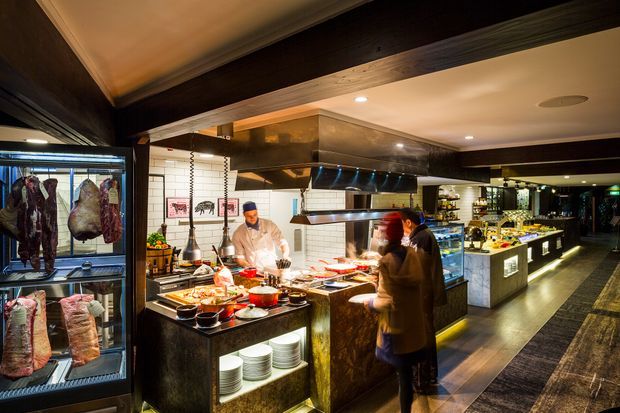 A warmly lit open restaurant kitchen and dining-service area, with chefs preparing food at a central cooking counter, dark stone-look counters, white tiled walls, stainless-steel extraction canopy, illuminated plate storage, refrigerated meat display, and a bar or service counter extending into the background. Exposed dark timber beams and low ceilings create a dramatic, intimate commercial interior.
