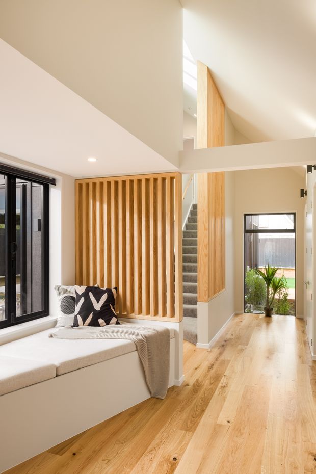 Bright contemporary residential hallway and stair landing with a double-height void. The space features pale timber flooring, a built-in window seat, a vertical timber batten screen enclosing the stair, white walls, black-framed glazing, and carpeted stairs. Natural light enters through the high-level opening and windows, creating an airy, warm interior.